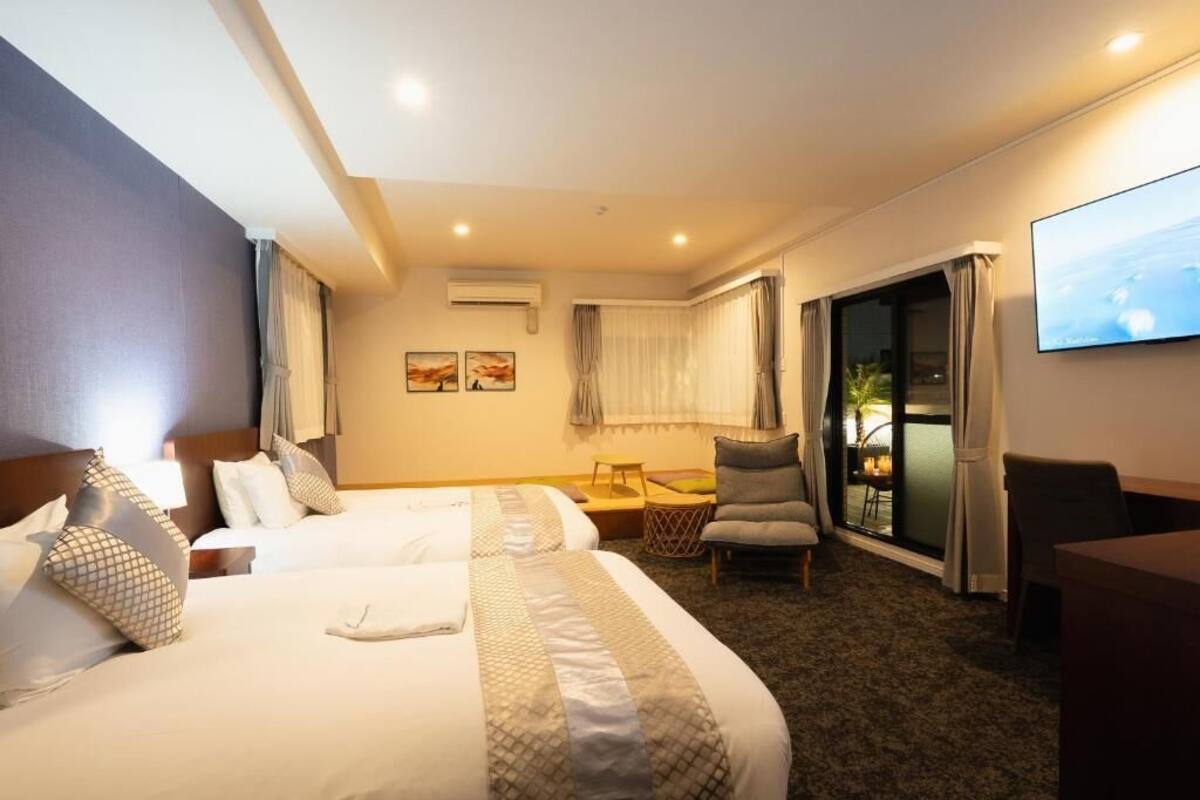 A hotel room with two beds, a desk, a chair, a TV, and a balcony with curtains.