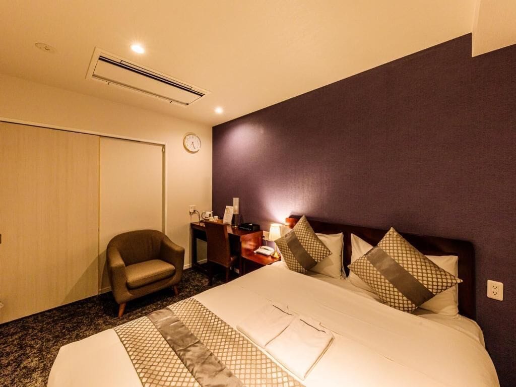 A hotel room with a large bed, a chair, a desk with a lamp, and a clock on the wall.