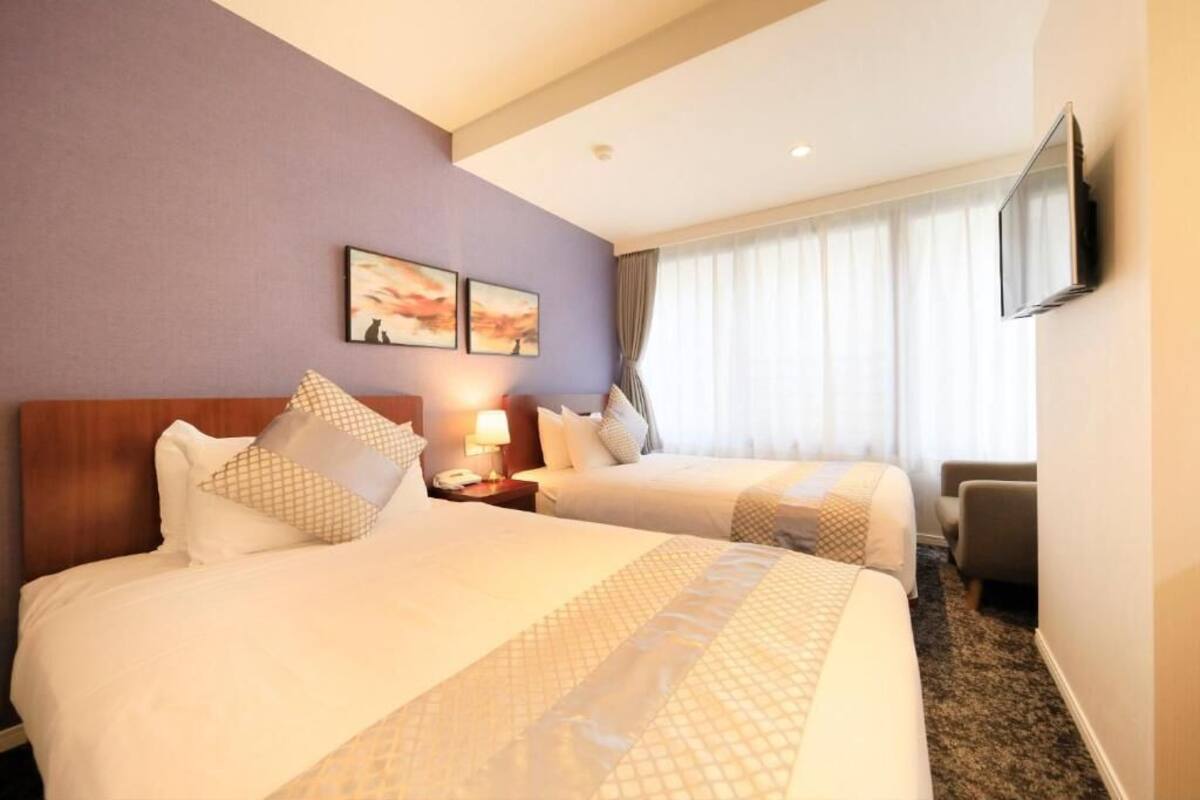 A hotel room with two beds, a flat-screen TV mounted on the wall, and a window with curtains.
