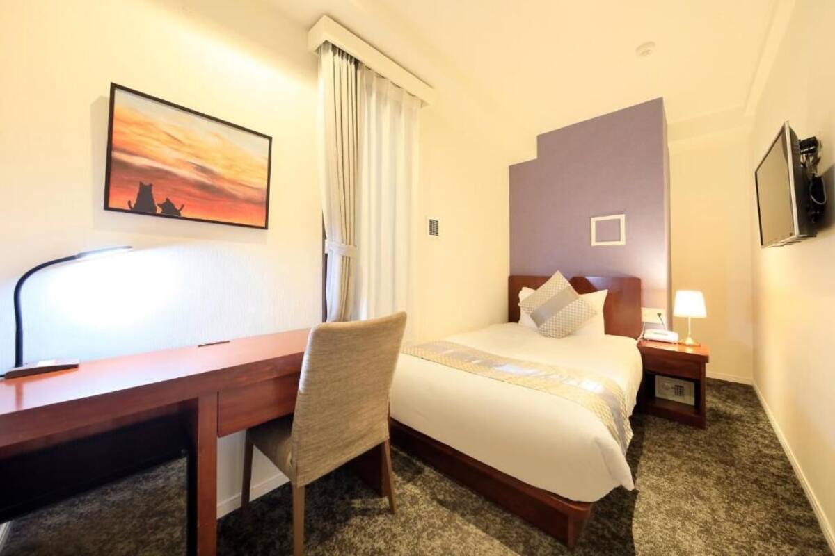 A hotel room with a bed, a desk, a chair, a nightstand, a TV, and a painting on the wall.