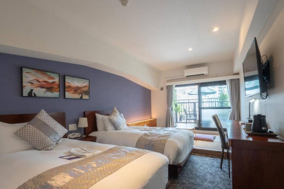 A hotel room with two beds, a desk, a chair, a flat-screen TV, and artwork on the wall.