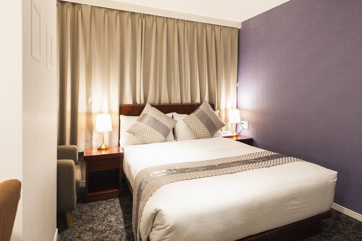 A hotel room with a bed, two bedside tables with lamps, a chair, and a large window with curtains.
