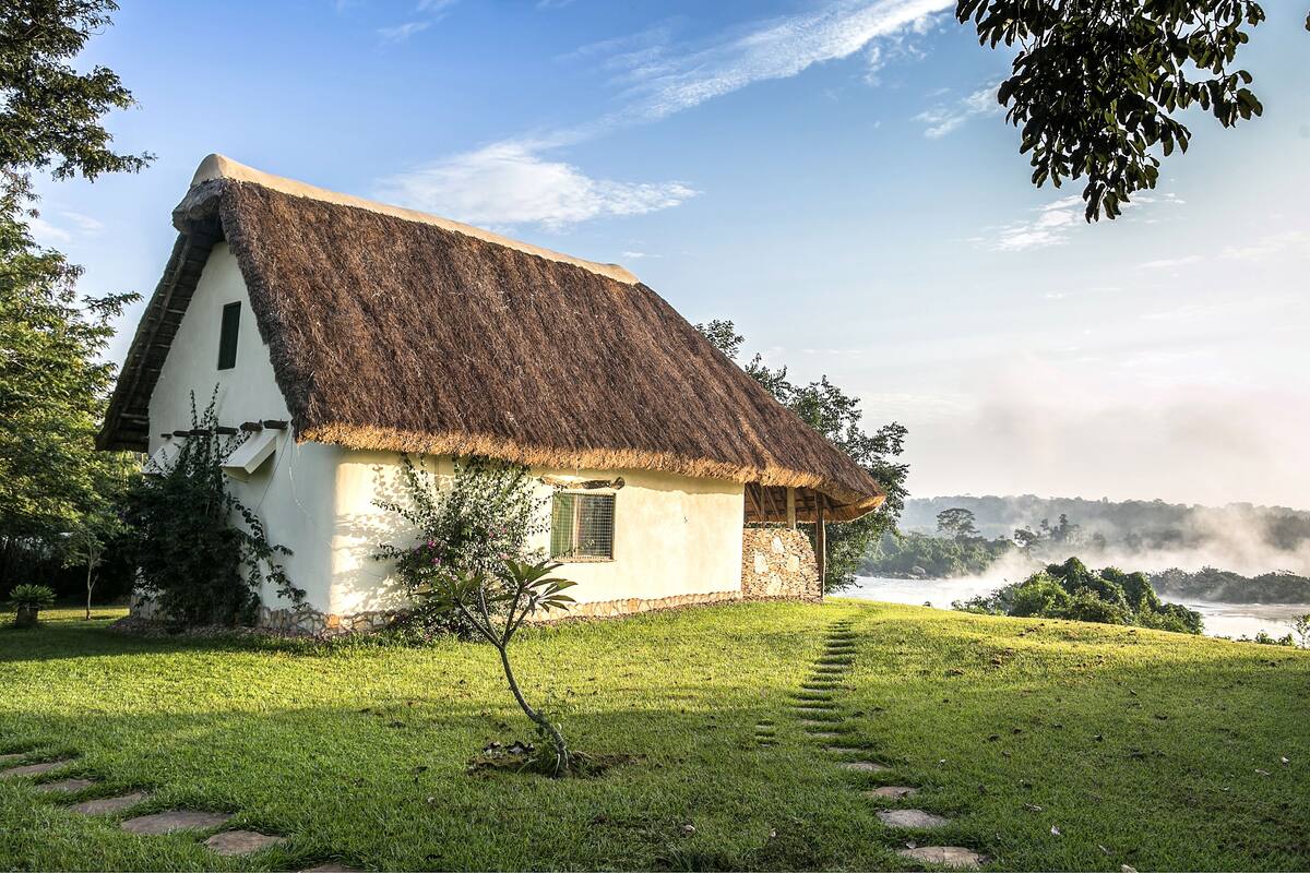 Una casa con tetto di paglia e una piccola finestra, immersa nel verde e con un sentiero che conduce all'acqua.
