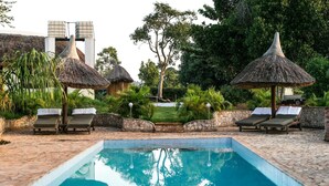 Una piscina con sillones de descanso y sombrillas de paja, rodeada de vegetación y muros de piedra.