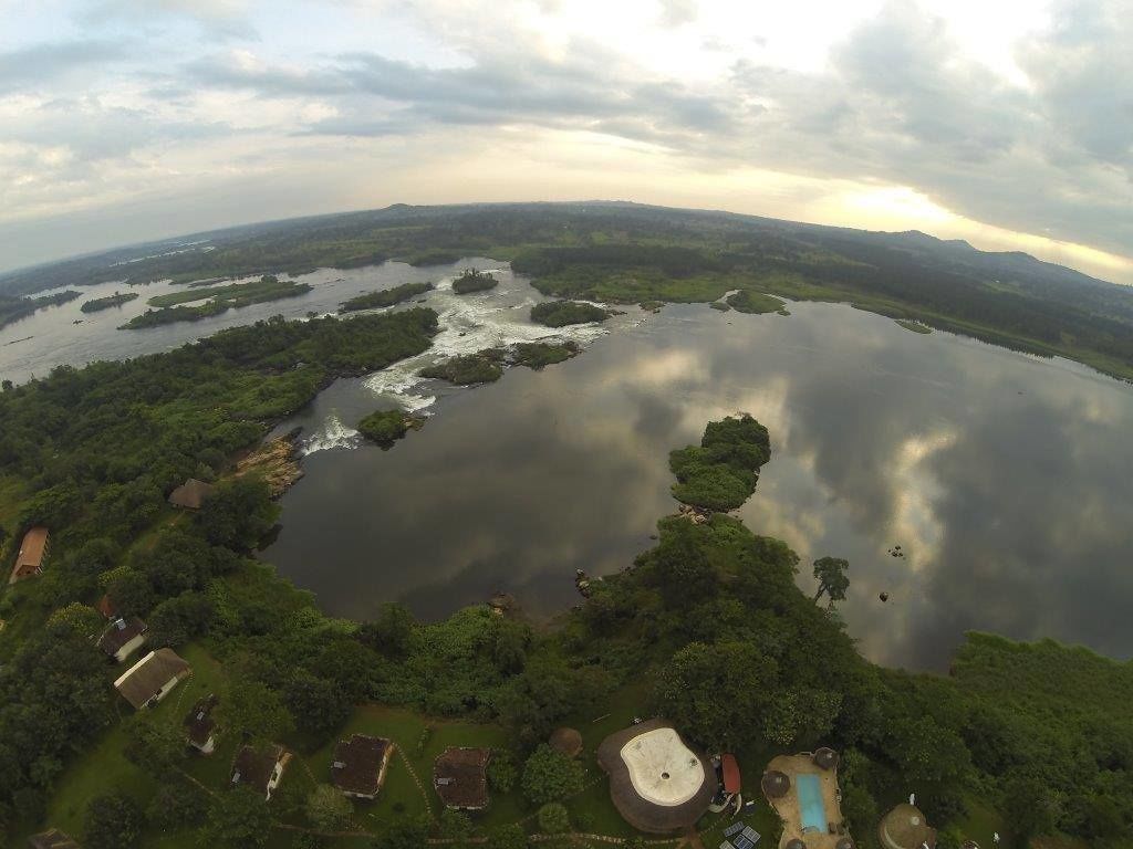 An aerial view of a lake surrounded by dense greenery and small buildings, with a cloudy sky overhead.