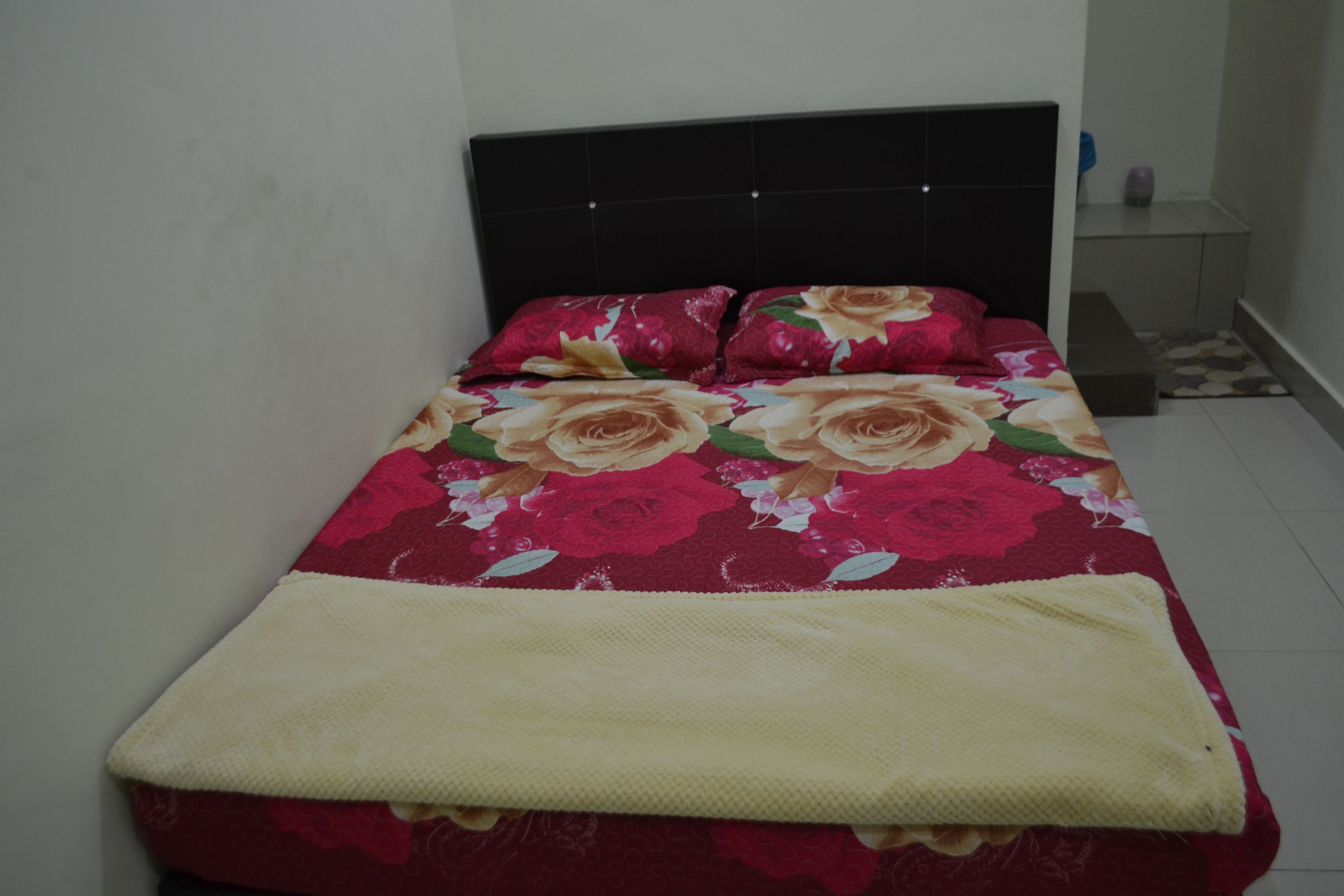 Una cama con una colcha estampada y dos almohadas, una mesita de noche con una lámpara, y un pequeño banco.
