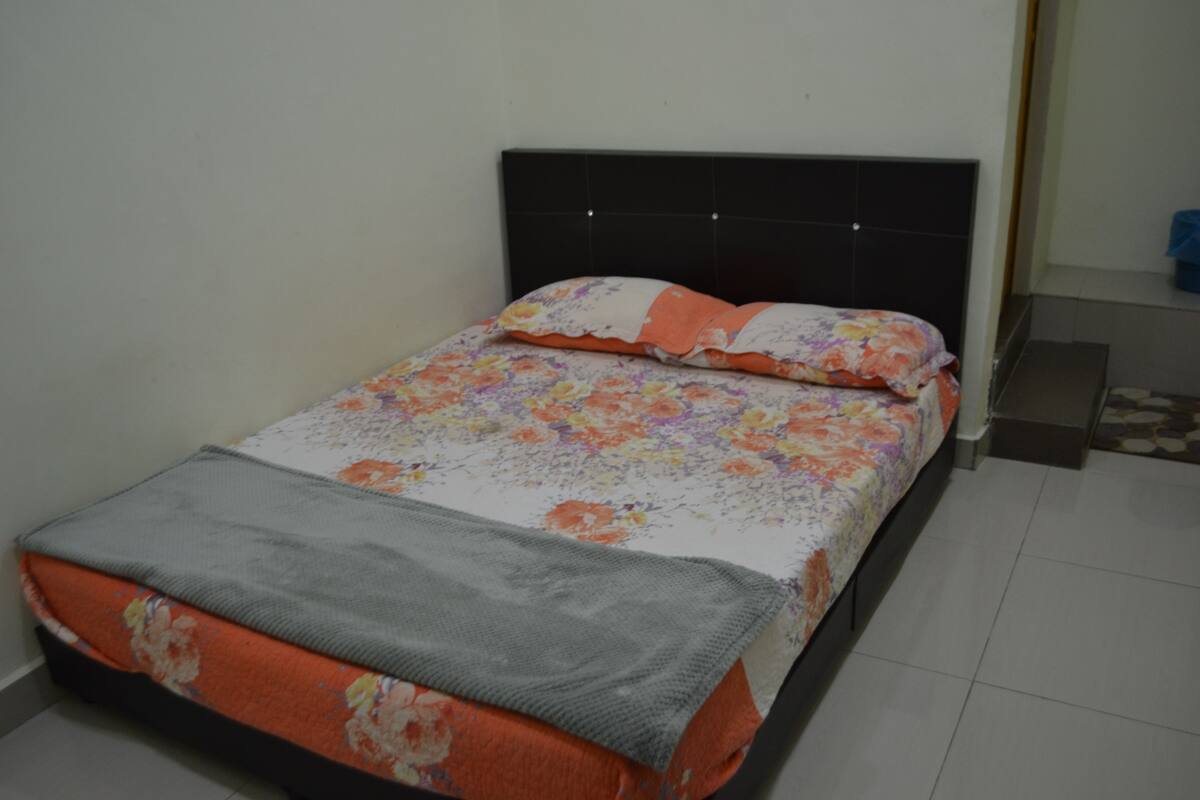 Una cama con colcha estampada, manta gris, cabecera negra y suelo de baldosas.