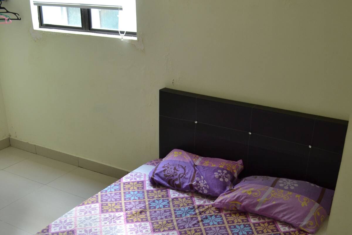 Una cama con una colcha estampada y dos almohadas, una ventana con persianas y una pared lisa.