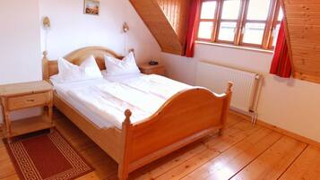 Ein Schlafzimmer mit einem hölzernen Bett, einem Nachttisch, einem roten Teppich, einem Fenster mit roten Vorhängen und einer Holzbalkendecke.