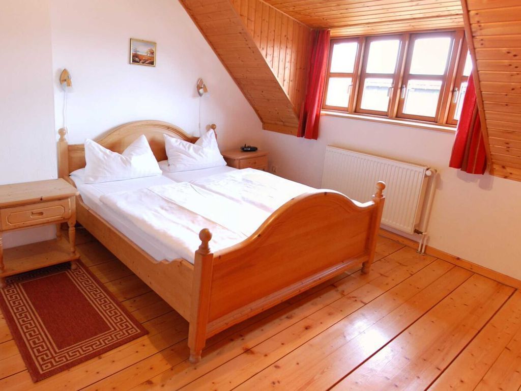 Ein Schlafzimmer mit einem hölzernen Bett, einem Nachttisch, einer Kommode, einem Heizkörper und einem Fenster mit roten Vorhängen.