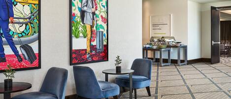 A hotel lobby with blue chairs, a small table with a plant, and a painting of a man with a suitcase.