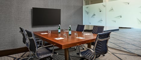 A conference room with a wooden table, six chairs, a flat-screen TV, and a glass partition with geometric patterns.