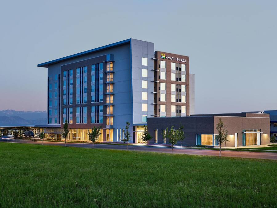 A modern hotel with a prominent 'Hyatt Place' sign, featuring a mix of glass and brick facades, and a parking lot in the foreground.