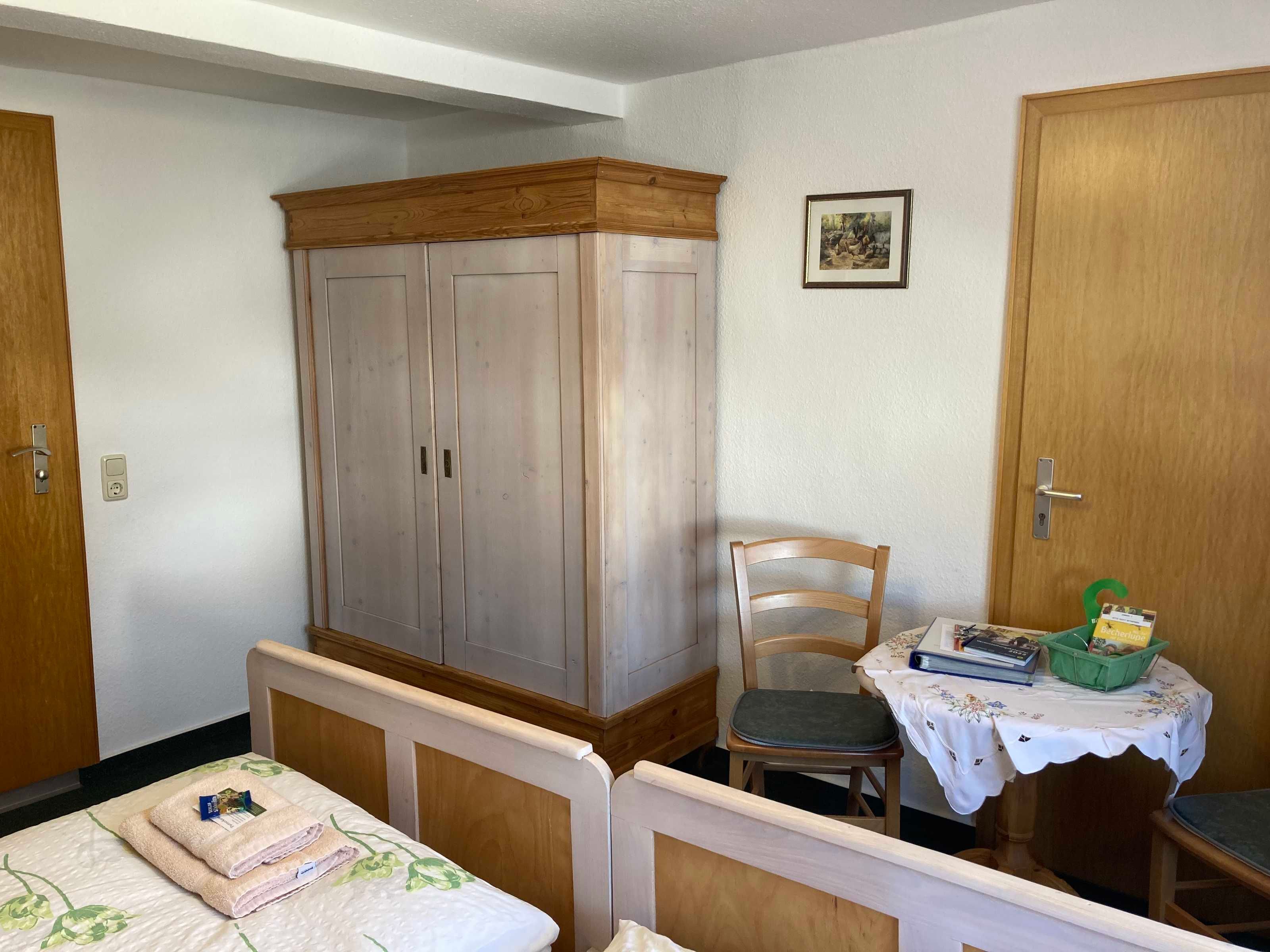 A bedroom with a bed, a wooden wardrobe, a chair, a small table with a basket, and a framed picture on the wall.