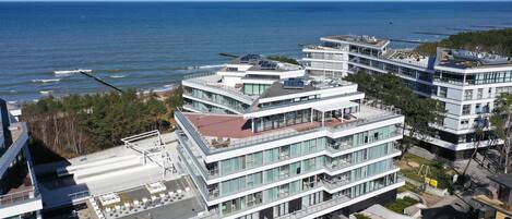A modern multi-story hotel with balconies overlooking the sea, featuring a rooftop terrace and a landscaped courtyard.