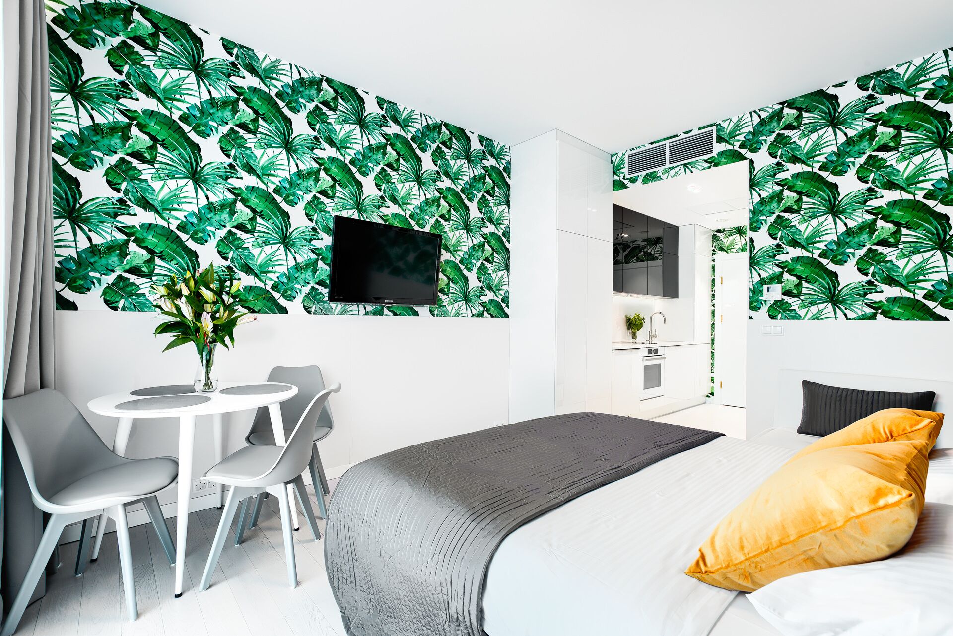 Een moderne slaapkamer met behang met een tropisch bladmotief, een bed met een grijs hoofdeinde, een geel kussen en een kleine ronde tafel met stoelen.