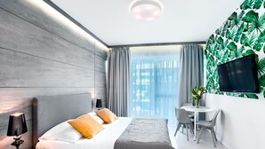 A modern hotel room with a large bed, a bedside table with a lamp, a small desk with a chair, a TV mounted on the wall, and a window with curtains. - Apartamenty Sun & Snow Dune B (Mielno)