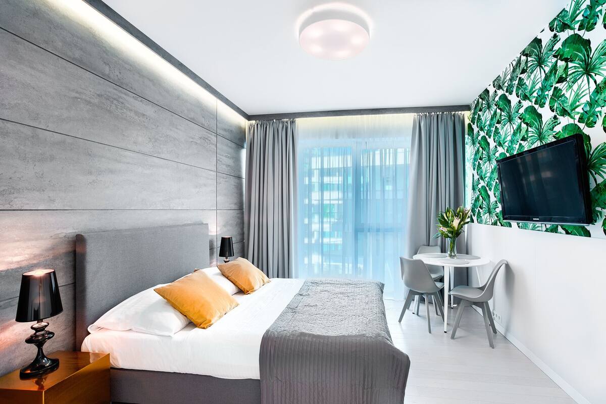 A modern hotel room with a large bed, a bedside table with a lamp, a small desk with a chair, a TV mounted on the wall, and a window with curtains.