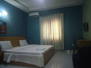 Quarto de hotel com cama, mesa de cabeceira, aparelho de ar condicionado, janela com cortinas e um quadro na parede.