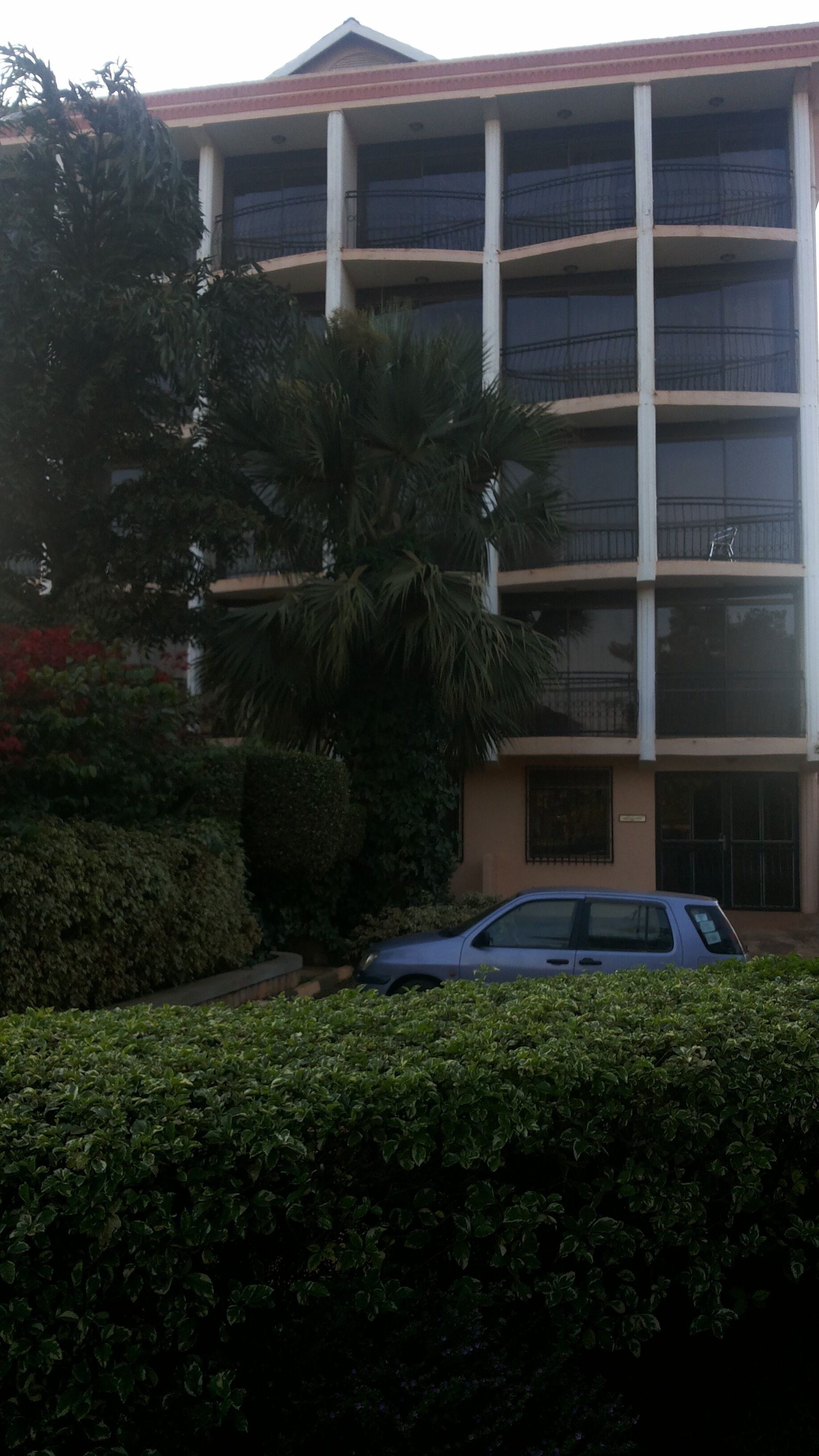 A multi-story building with balconies, surrounded by dense greenery and a parked car.
