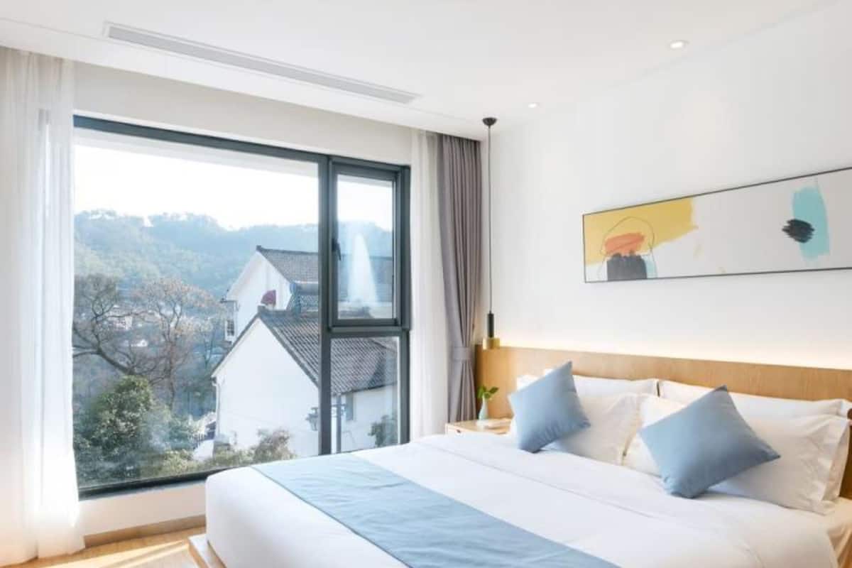 Un dormitorio moderno con una cama grande, vista de una casa a través de una ventana y un cuadro en la pared.
