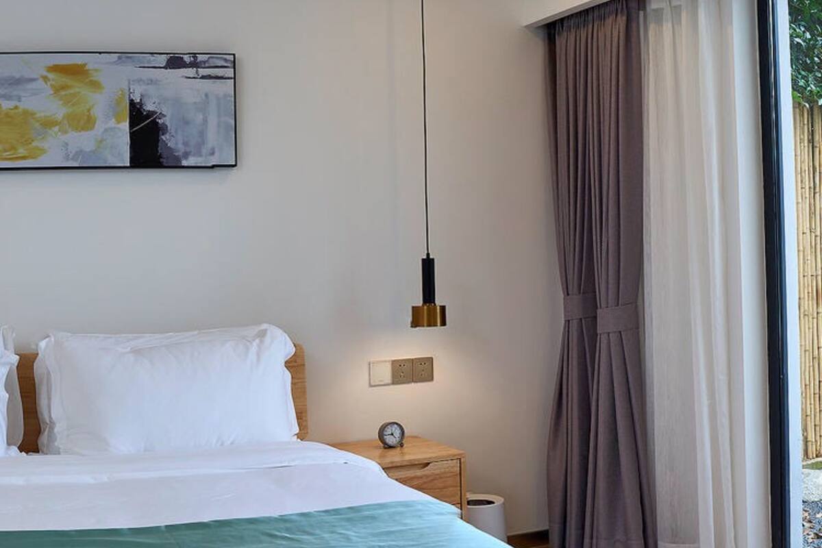 Una cama bien hecha con sábanas blancas, una manta verde, una mesita de noche con un reloj y una lámpara colgante moderna.