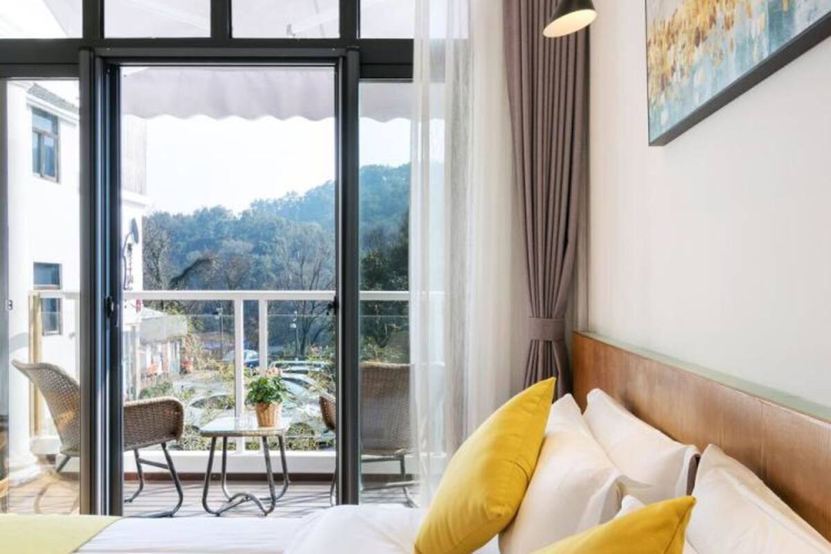 Una habitación de hotel con una cama grande, dos almohadas amarillas, una mesita de noche, una lámpara, una ventana que ofrece vistas a un balcón y árboles, y un cuadro en la pared.