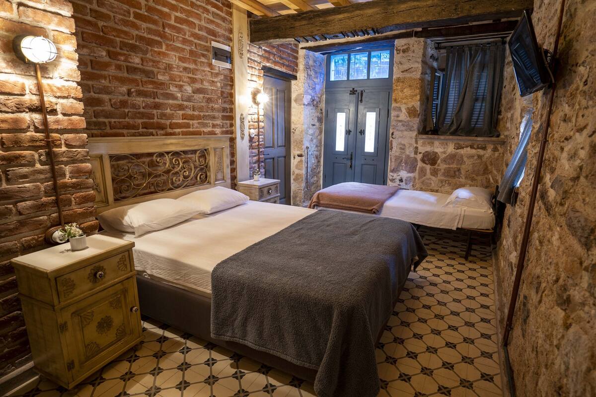 A rustic room with a brick wall, a wooden bed with a headboard, a bedside table with a lamp, and a small wooden dresser.