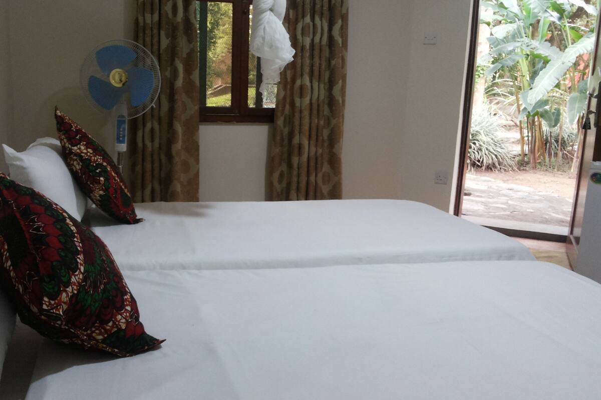 A hotel room with two beds, a standing fan, and a view of the outdoors through a window.