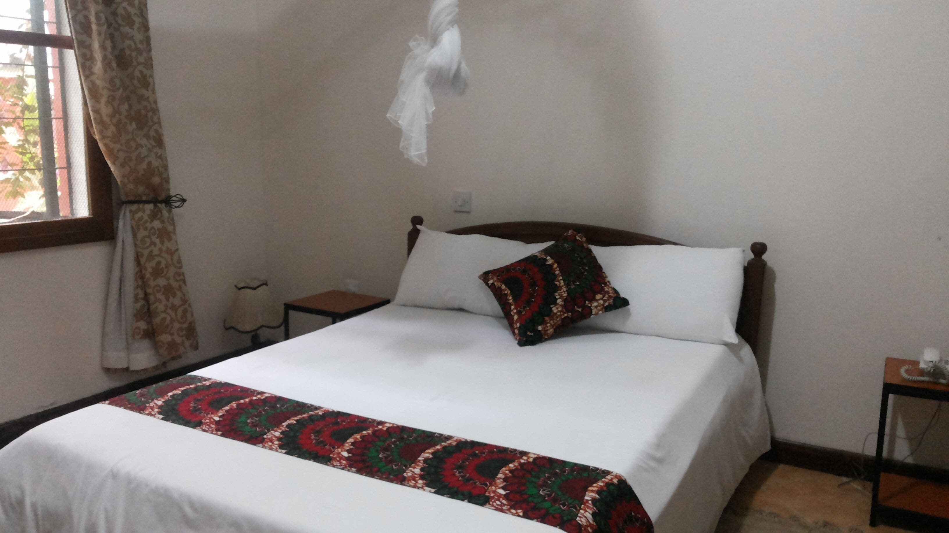 Un llit amb estriat blanc i una manta colorida, un capçal de fusta, una taula de nit amb una làmpada, i una finestra amb cortines.