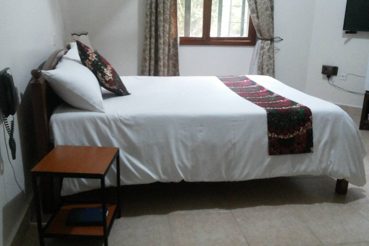 A neatly made bed with a white bedspread and a decorative blanket, a wooden nightstand with a book, a telephone, and a window with curtains.