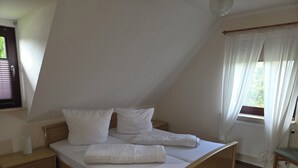 A bedroom with a sloped ceiling, a wooden bed frame, white bedding, a bedside table, and a window with curtains. - Upper apartment in Westeraccum with balcony (Westeraccum)