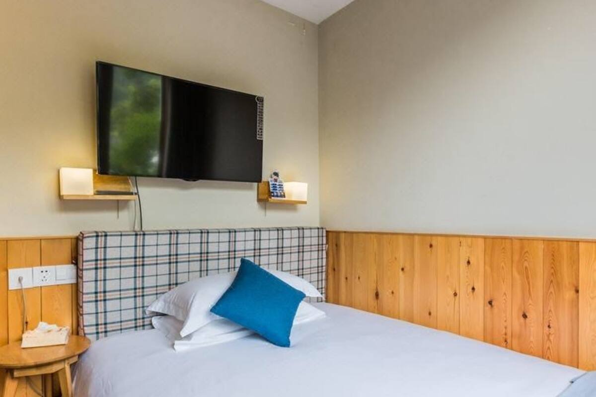 Una habitación de hotel con cama, televisión montada en la pared, mesita de noche y velador.