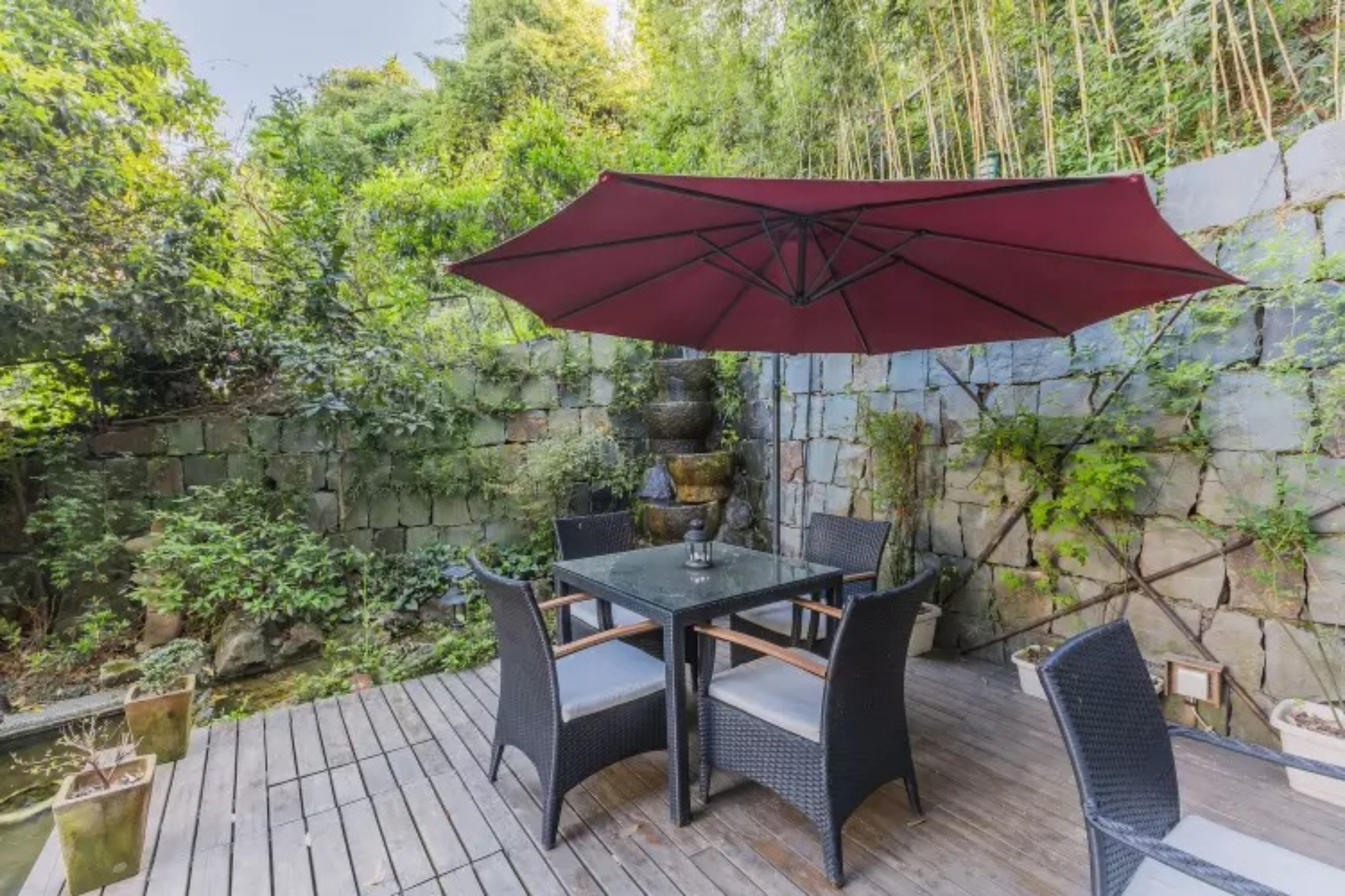 A patio with a table and chairs, a red umbrella, and a stone wall with greenery.