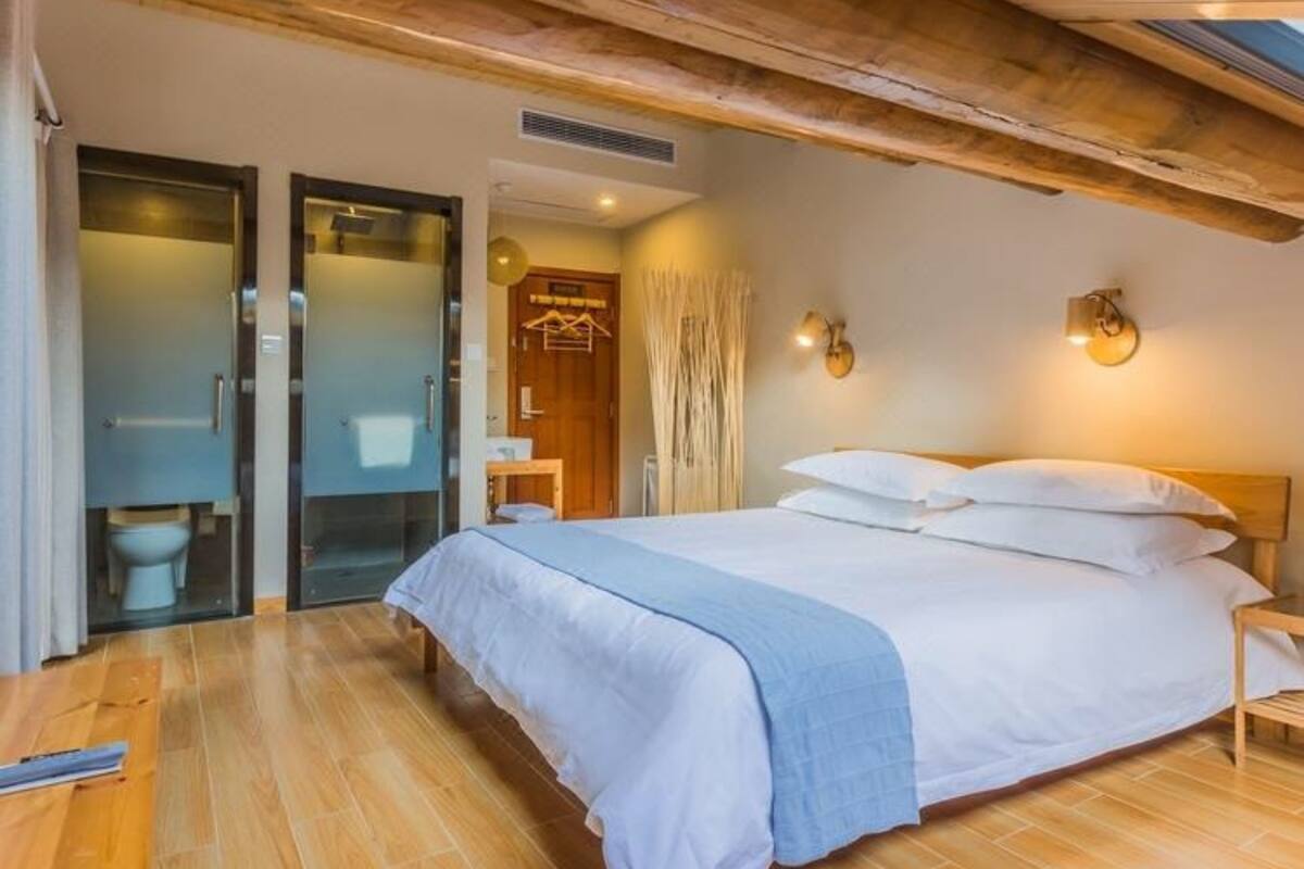 Un dormitorio con techo de madera, una cama grande con ropa blanca, suelo de madera y un baño con puerta de cristal.