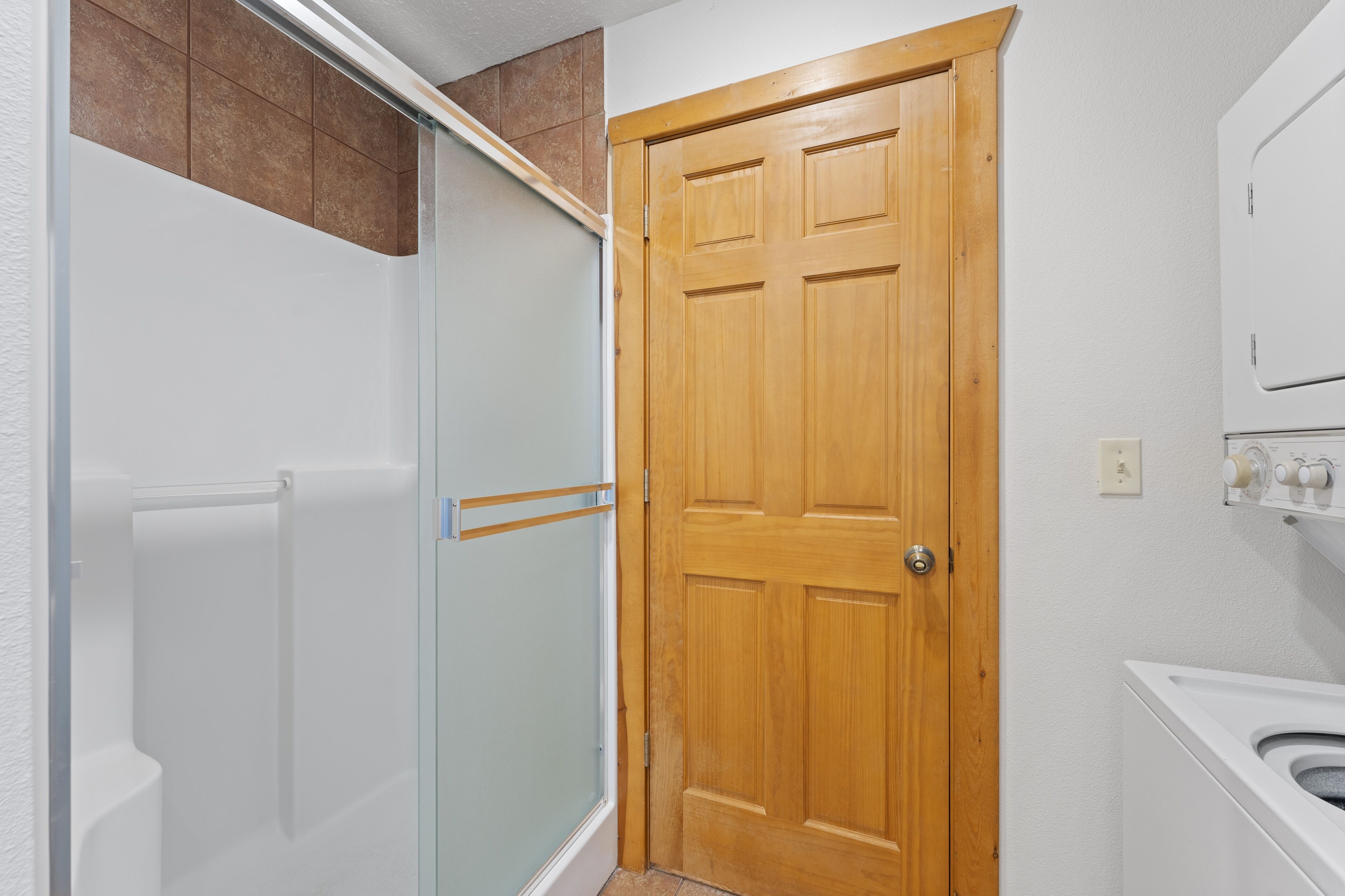 Un baño con puerta de madera, cabina de ducha de vidrio y lavadora blanca.