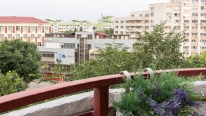 Un balcon avec une balustrade rouge, un pot de fleurs contenant des fleurs violettes et, en arrière-plan, une vue sur des immeubles à plusieurs étages.