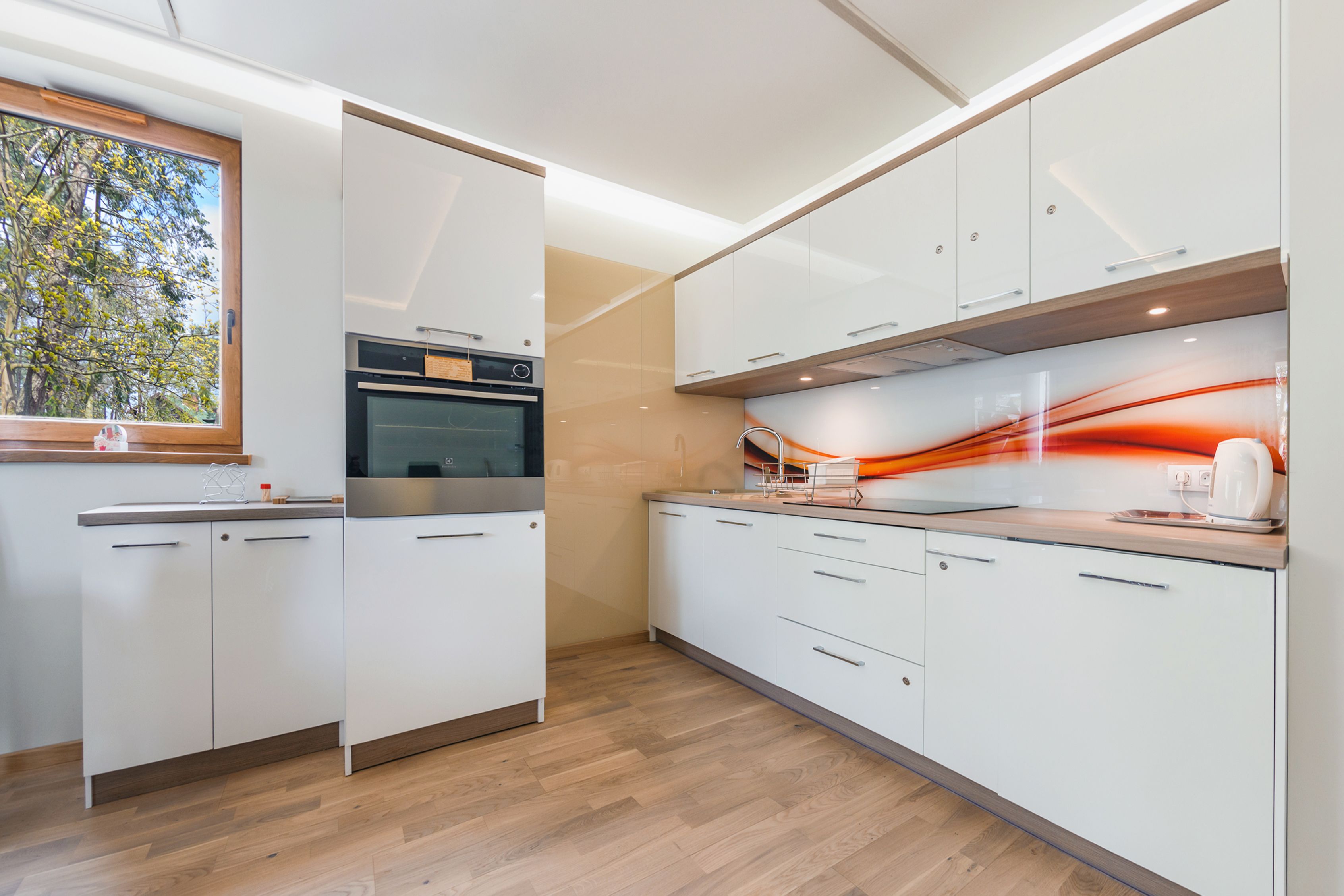 A modern kitchen with white cabinetry, a built-in oven, and a window with a view of trees.