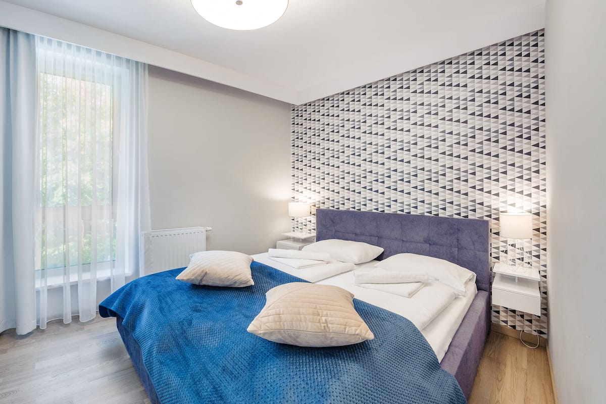 Una habitación de hotel moderna con una cama grande, un cabecero con estampado geométrico, una ventana con cortinas translúcidas y una mesita de noche con lámpara.
