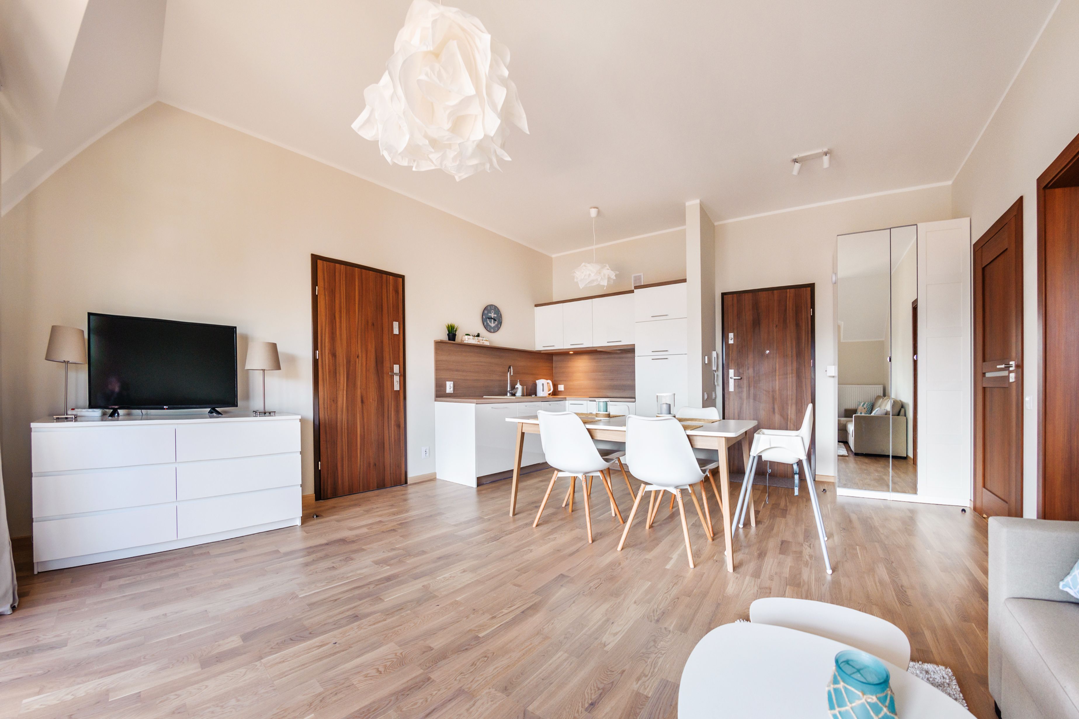 A modern living room with a flat-screen TV, a dining area with a table and chairs, and a kitchen with wooden cabinets and a white countertop.