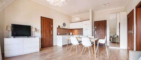 A modern living room with a flat-screen TV, a dining area with a table and chairs, and a kitchen with wooden cabinets and a white countertop.