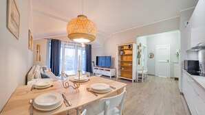 A modern kitchen with a built-in stove, a dining area with a wooden table set for four, and a living area with a TV and bookshelf. - Apartamenty Sun&Snow Rezydencja Park (Mielno)