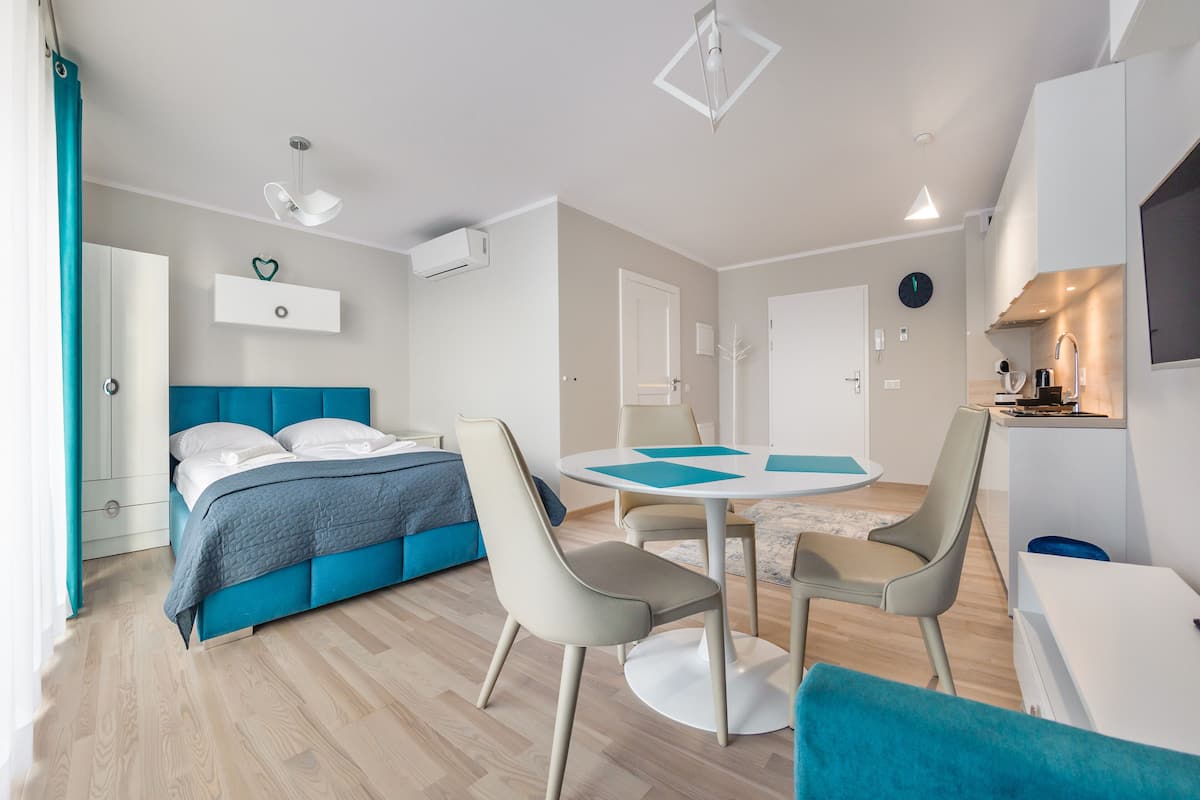 Un dormitorio moderno con una cama azul, una mesa de comedor redonda con sillas, una pequeña cocina y un televisor en la pared.