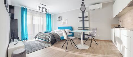 A modern hotel room with a bed, a desk, a chair, a small table, and a kitchenette.