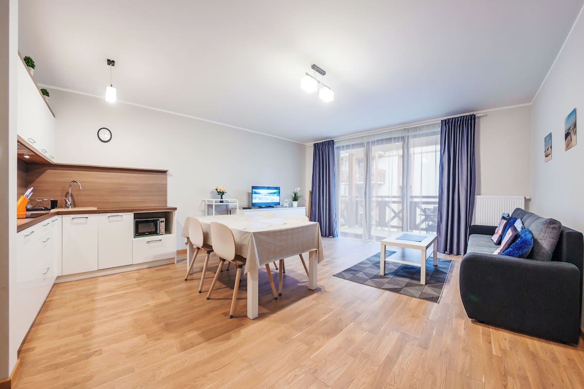 Un apartamento moderno con cocina, comedor y sala de estar. Cuenta con piso de madera, muebles de cocina blancos, una mesa de comedor con sillas, un sofá y un televisor.
