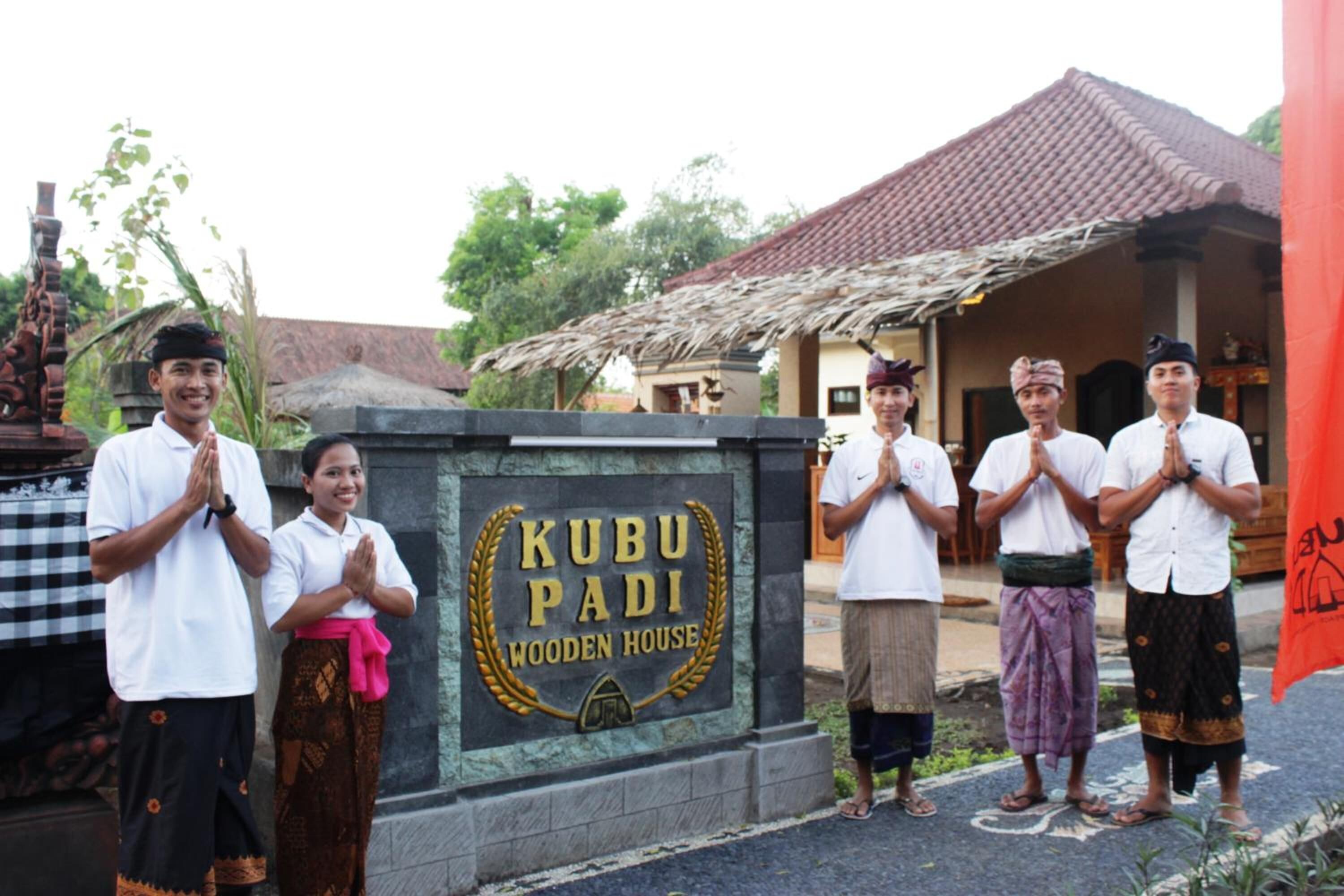 Un grupo de personas de pie frente a una casa de madera con un letrero que dice 'Kubu Padi Wooden House'.