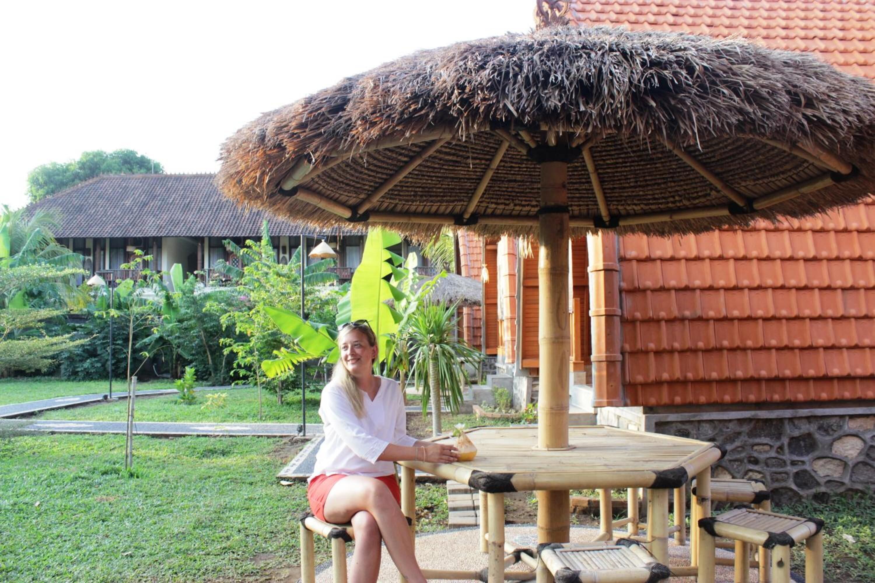 A person sitting at a round wooden table with a thatched umbrella, surrounded by greenery and a building with a tiled roof.