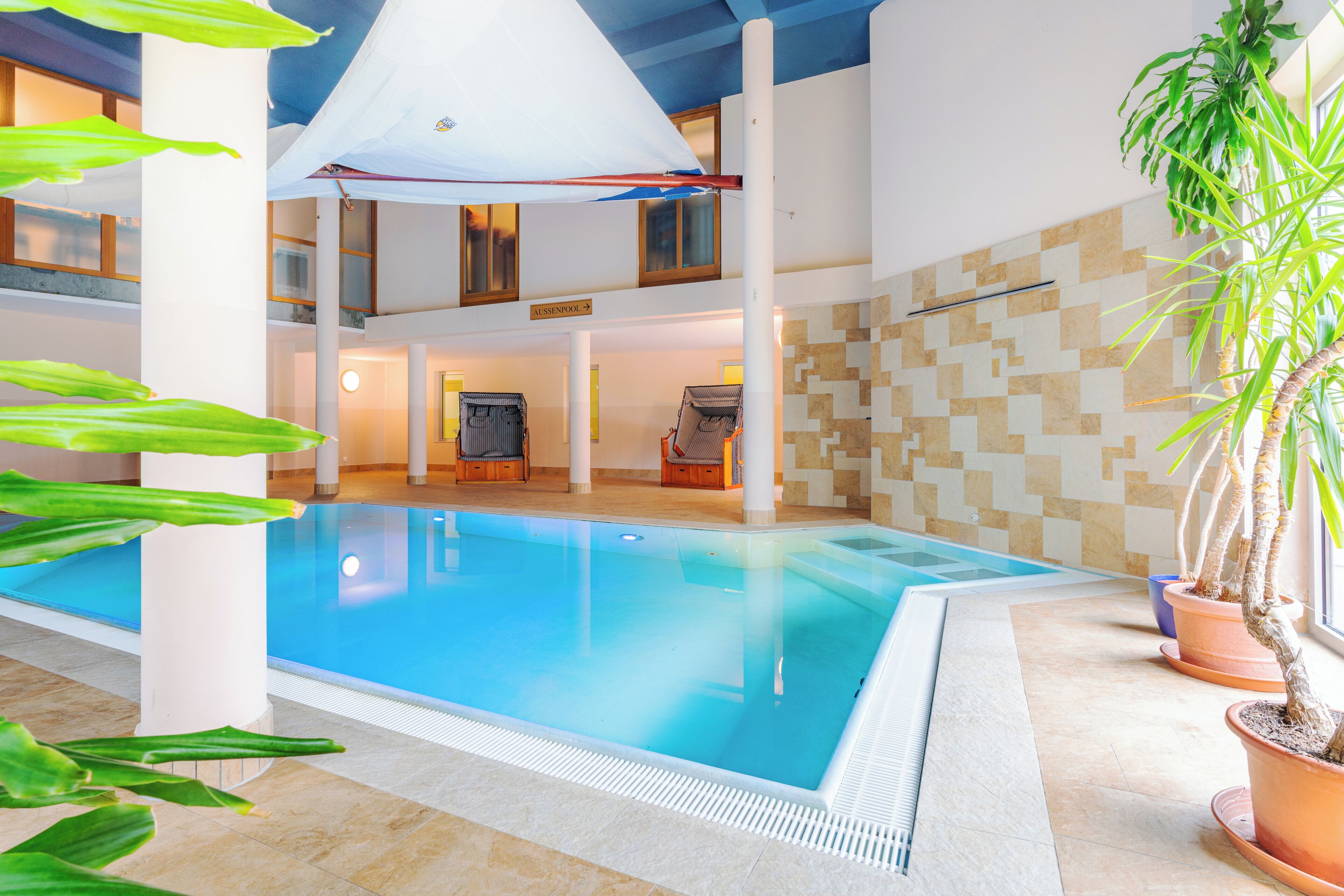 An indoor swimming pool with a tiled floor, a ceiling with a white canopy, and potted plants.