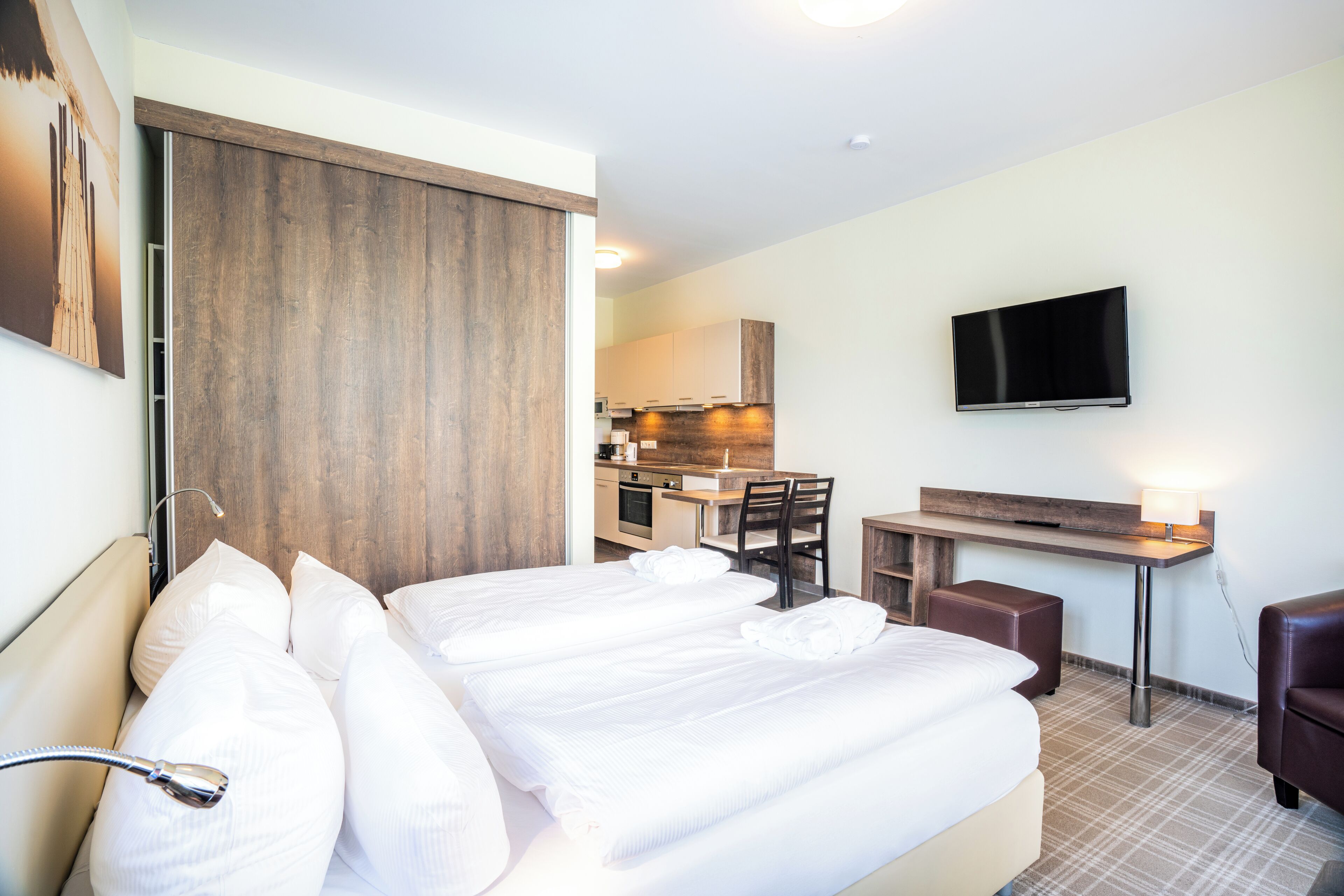 Ein Hotelzimmer mit einem Bett, einem Schreibtisch, einem Fernseher und einer Küchenzeile im Hintergrund.