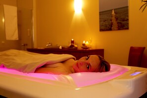 Uma pessoa deitada em uma cama com um aparelho de massagem, uma mesa de cabeceira com velas e um quadro na parede.